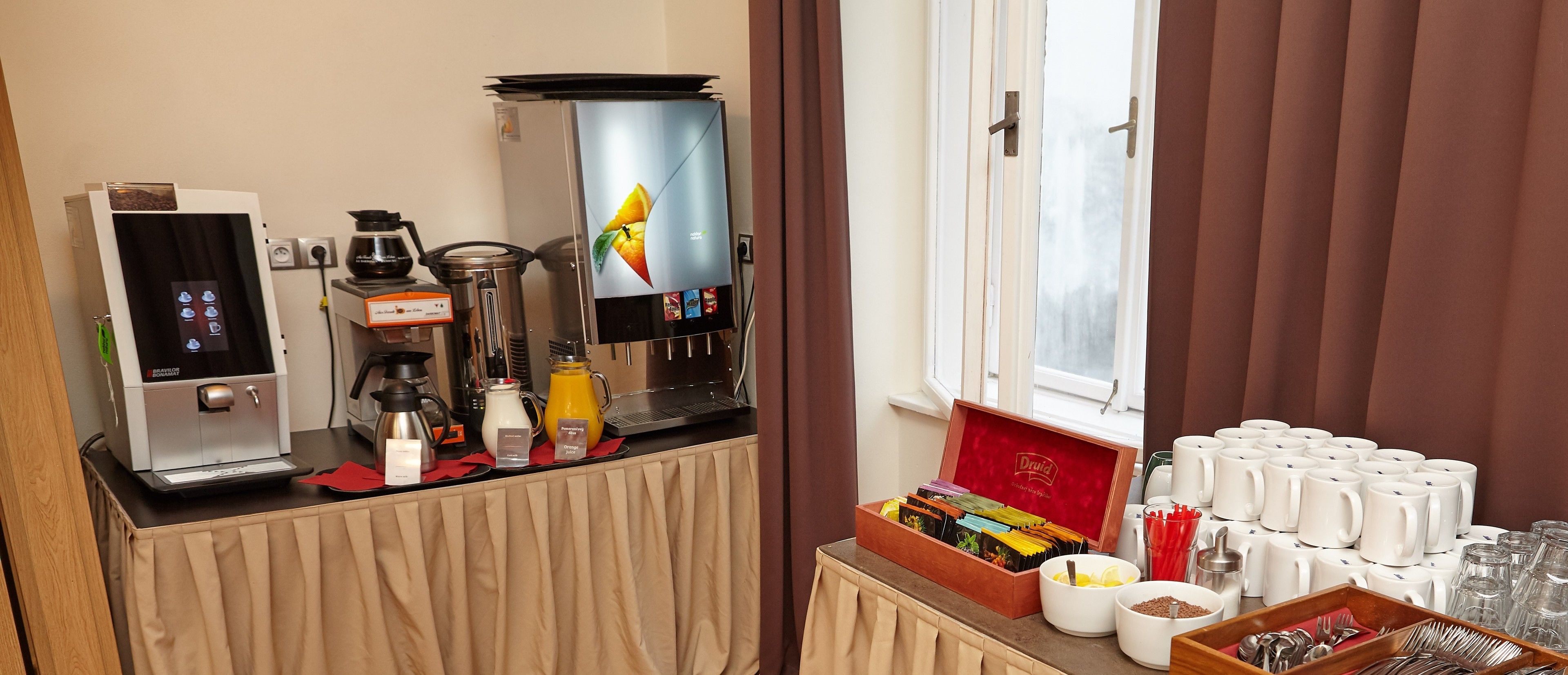 Quarto de hotel com espaço para café, geladeira, janela com cortinas e mesa com canecas e itens de café da manhã.