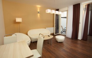Una habitación de hotel moderna con piso de madera, una mesa redonda, dos sillas blancas, un sofá y un pequeño reposapiés.