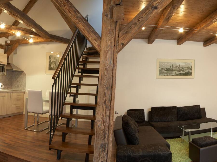 A modern interior with a wooden staircase, a kitchen with appliances, a living area with sofas and a TV, and a wooden ceiling.