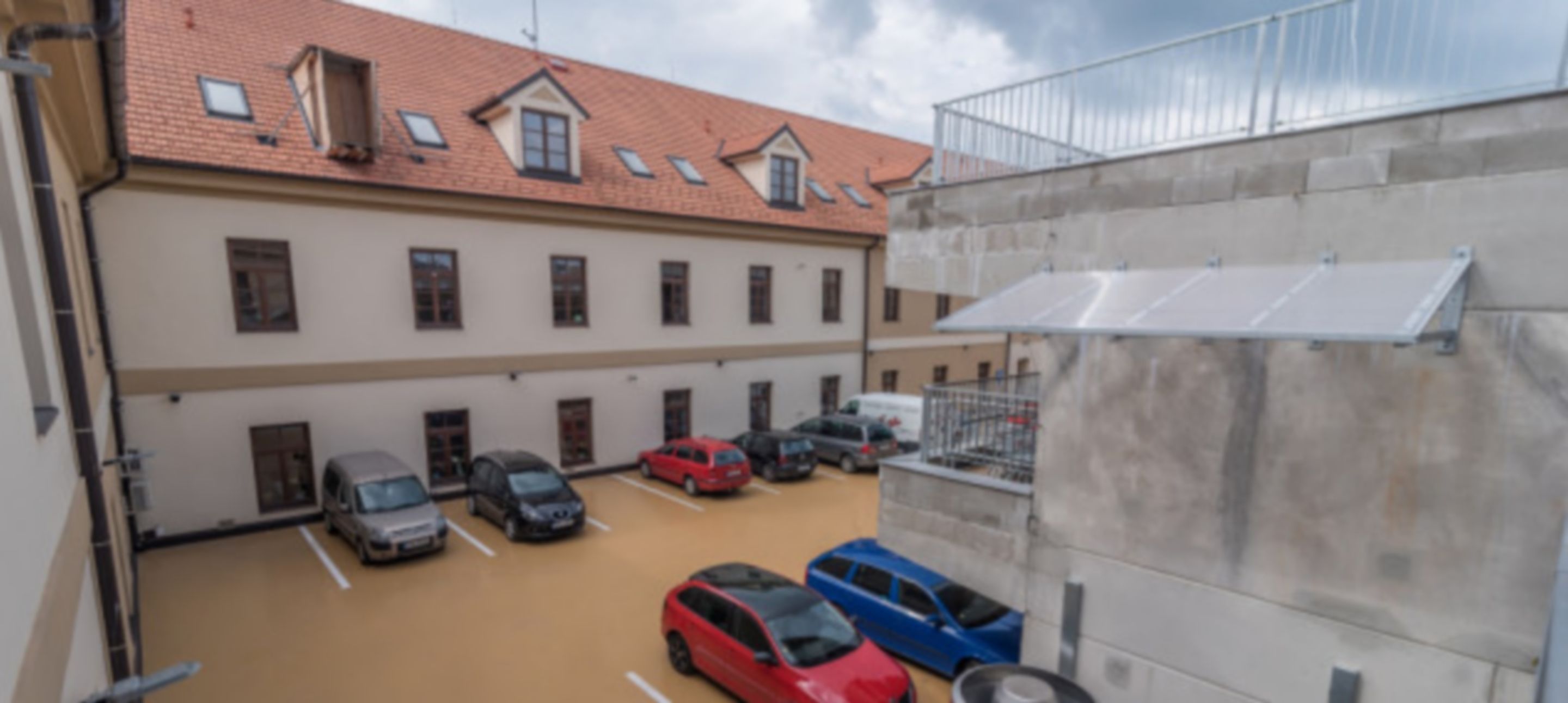 A parking area with multiple cars, a building with a tiled roof, and a concrete wall with a metal railing.