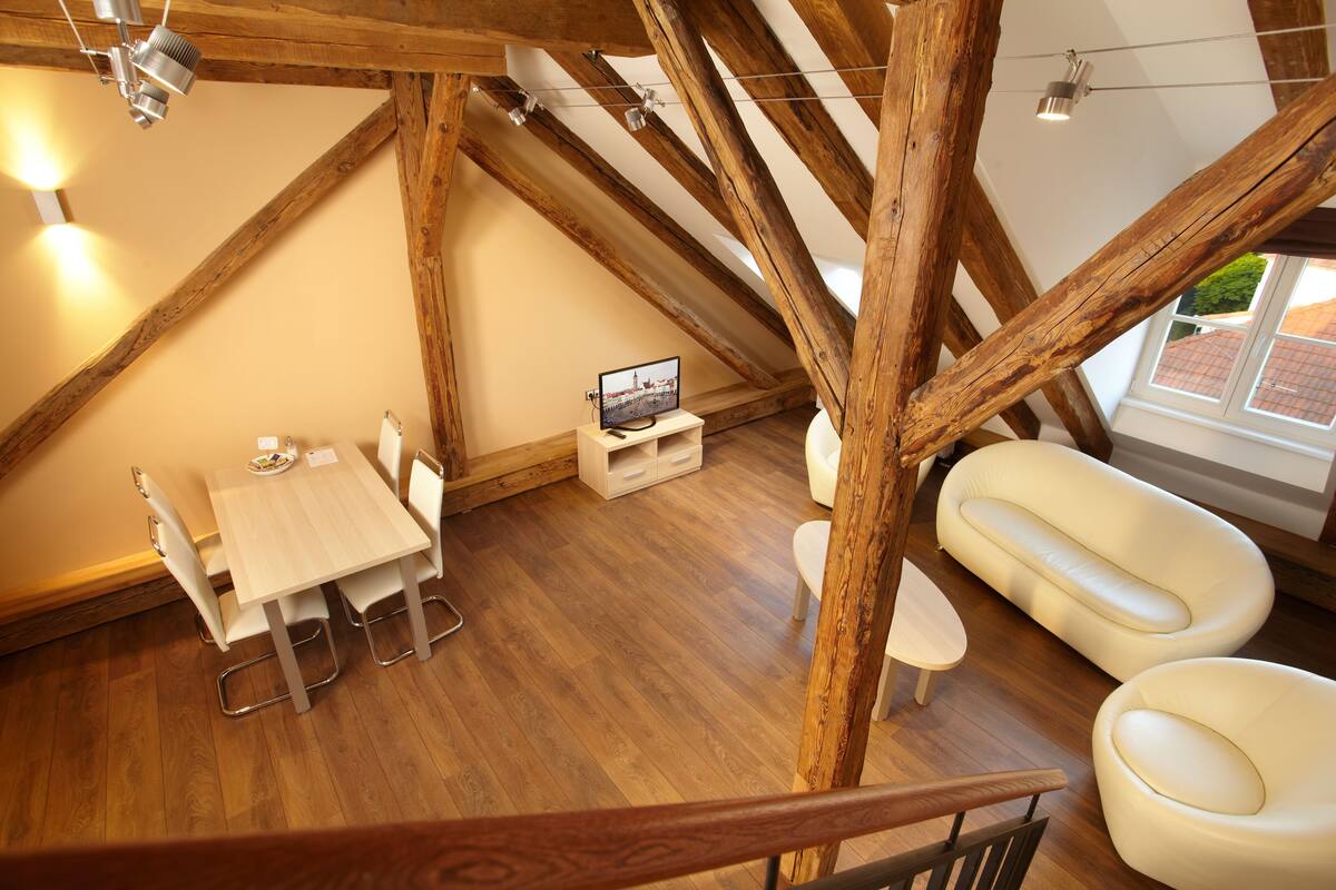 Una habitación estilo loft con vigas de madera a la vista, una zona de comedor con mesa y sillas, un sofá blanco y un televisor sobre un pequeño soporte.