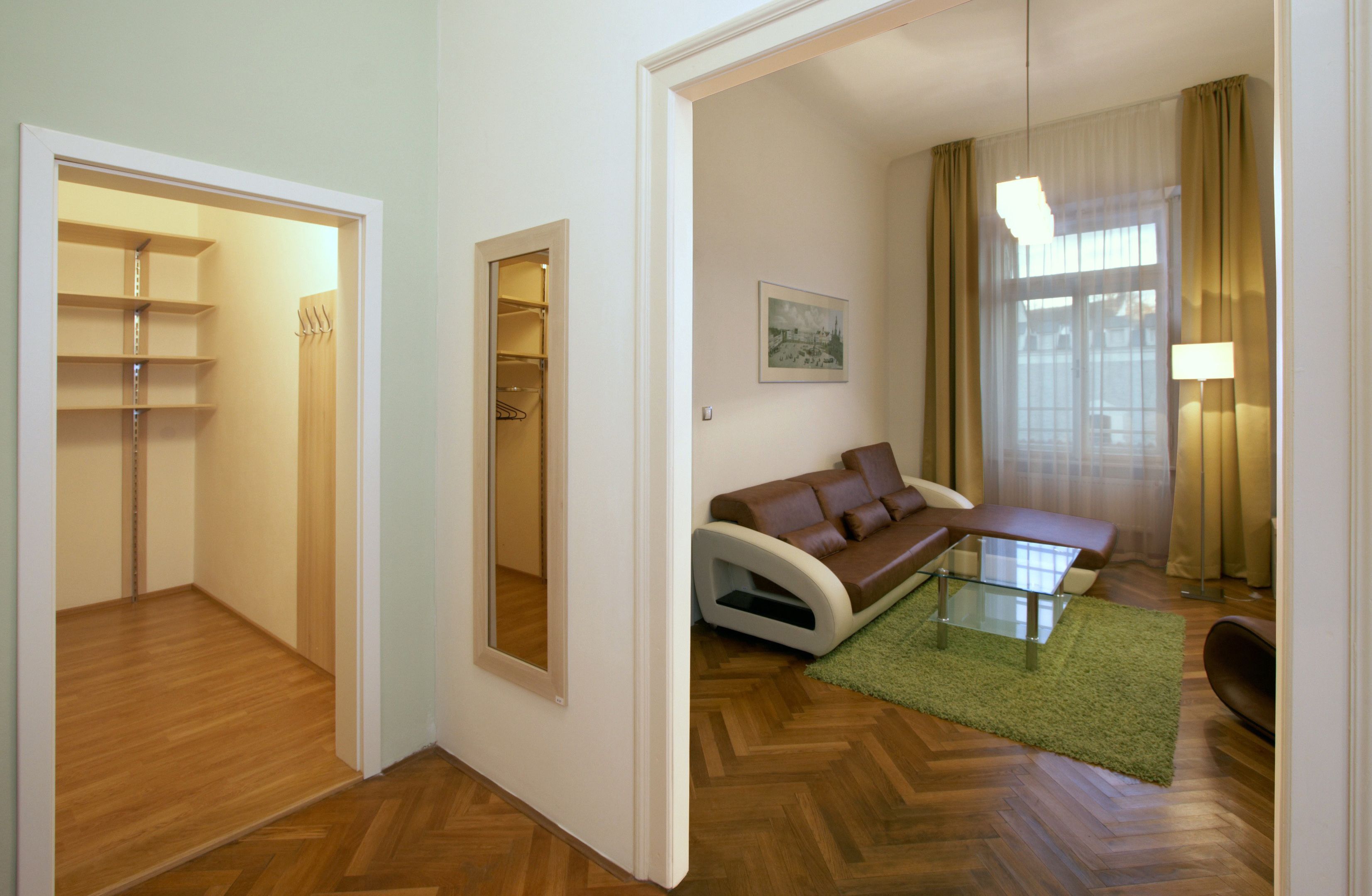 Ein modernes Wohnzimmer mit einem braunen Ledersofa, einem Glastisch und einem grünen Teppich. Holzfußboden und ein Flur mit einer weißen Tür zu einem Schrank.