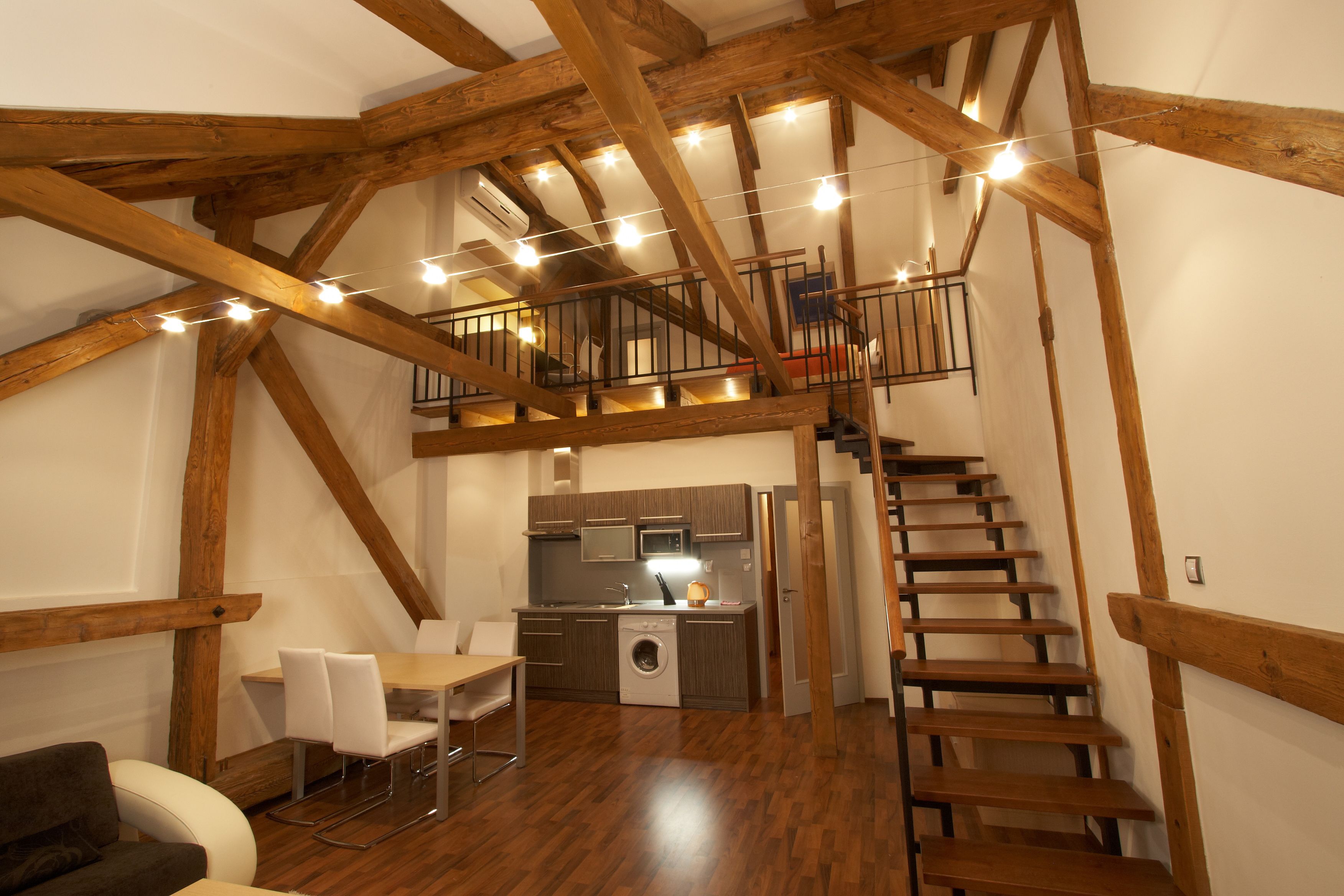 A spacious loft with exposed wooden beams, a kitchen area, a dining table with chairs, and a staircase leading to an upper level.