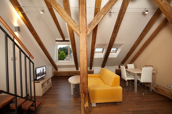 Un espacio tipo loft con vigas de madera a la vista, un sofá amarillo, una mesa de comedor con sillas y un televisor sobre un mueble de madera.
