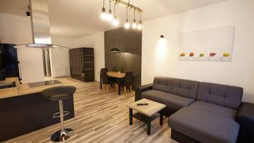 Ein modernes Wohnzimmer mit einem grauen Sofa, einem Holztisch und einem schwarzen Barhocker. Es gibt einen Essbereich mit Tisch und Stühlen sowie eine Küchenzeile mit Herd und Schränken.