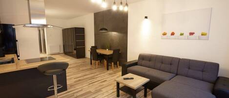 Una sala de estar moderna con un sofá gris, una mesa de centro de madera y un taburete negro. Hay una zona de comedor con mesa y sillas, y una cocina con estufa y armarios.
