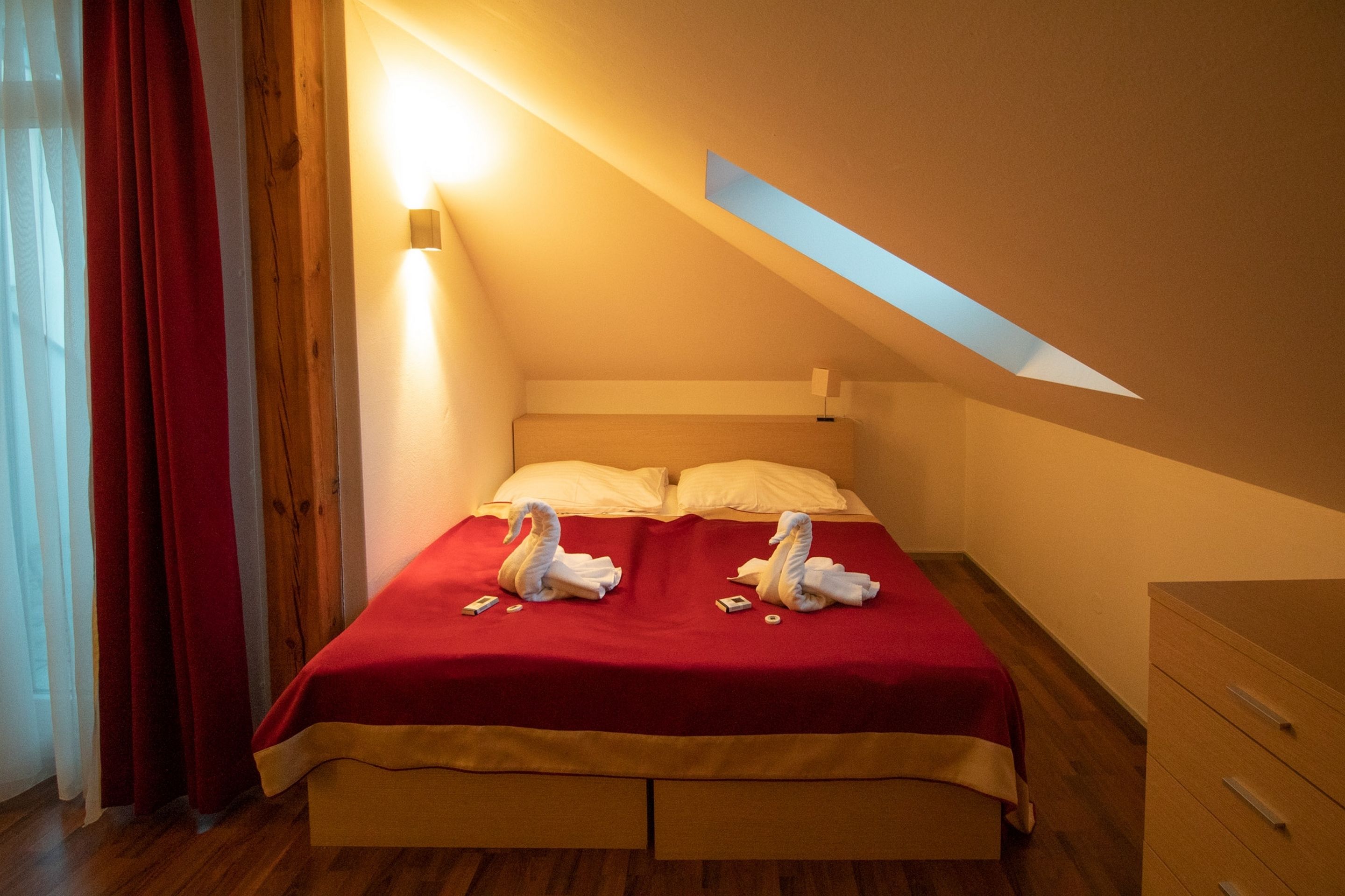 Un dormitorio con una colcha roja, dos toallas con forma de cisne, un cabecero de madera, una mesita de noche y una mesita auxiliar.