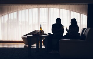 Una pareja sentada en un sofá en una habitación con poca luz, una ventana grande y una botella sobre una mesa.