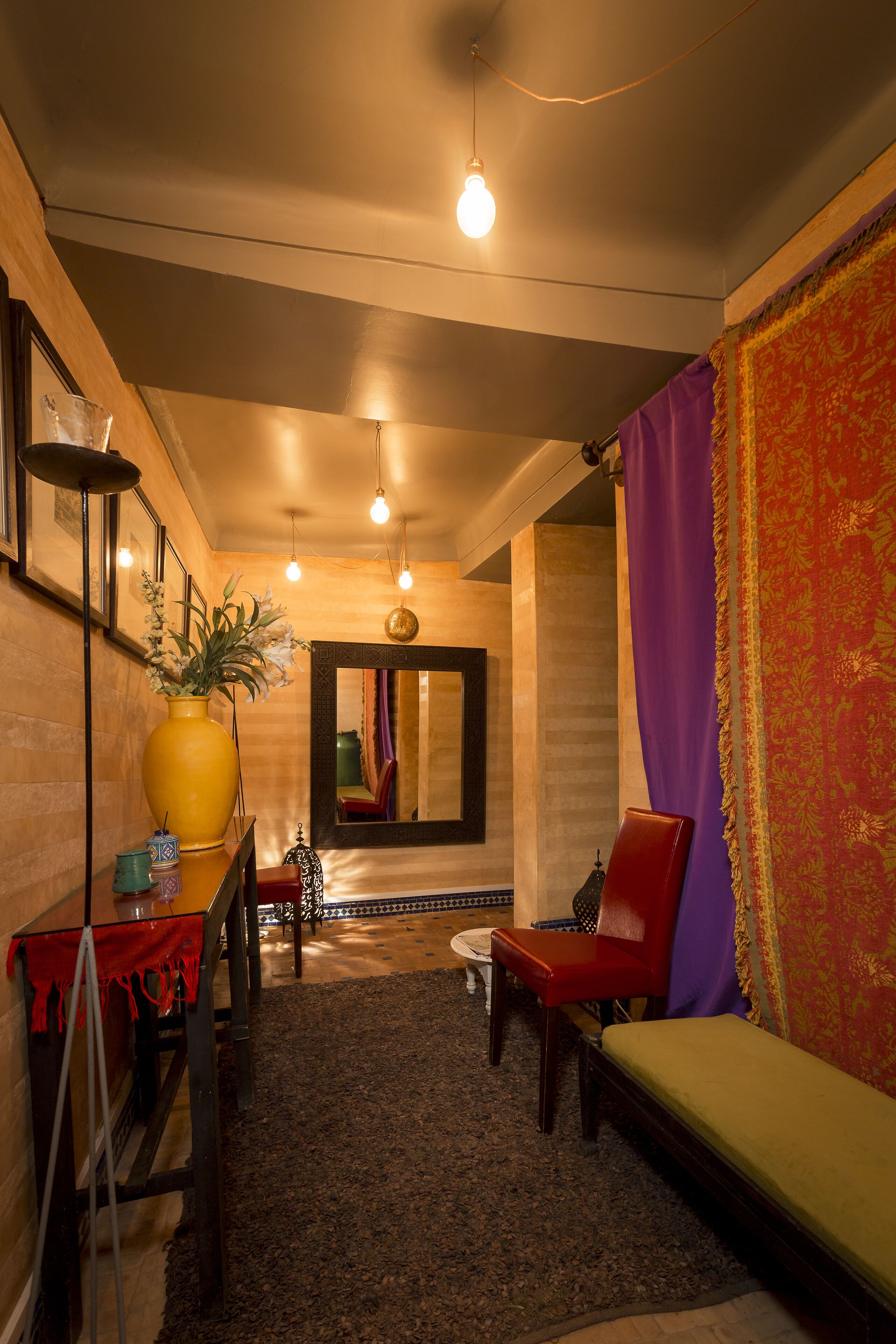 A room with a wooden floor, a large yellow vase with flowers, a red chair, a small round table, and a mirror on the wall.