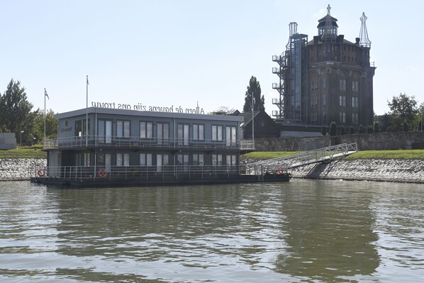 A riverboat with a flat roof and windows, docked near a stone embankment and a large building with a tower.