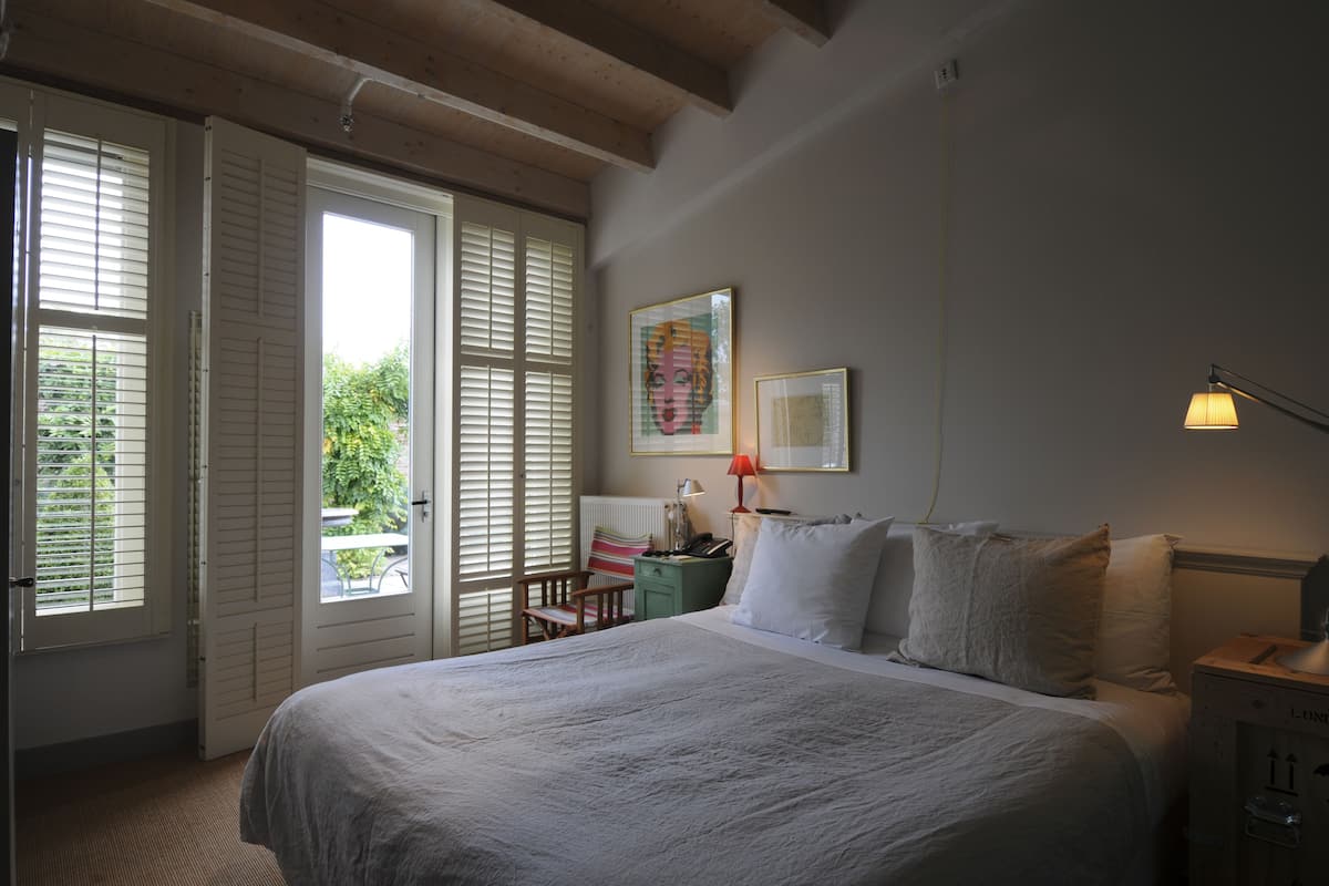 Un dormitorio con una cama grande, una mesita de noche, una silla, una ventana con contraventanas y un cuadro en la pared.