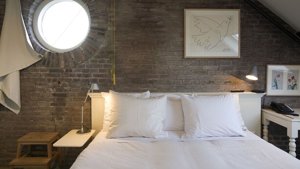 Un dormitorio con pared de ladrillo, un tragaluz circular, cama con ropa blanca, una lámpara de noche, un taburete de madera pequeño y un cuadro en la pared.