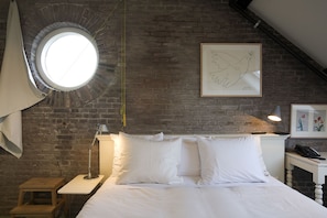 A bedroom with a brick wall, a round skylight, a bed with white bedding, a bedside lamp, a small wooden stool, and a framed picture on the wall.