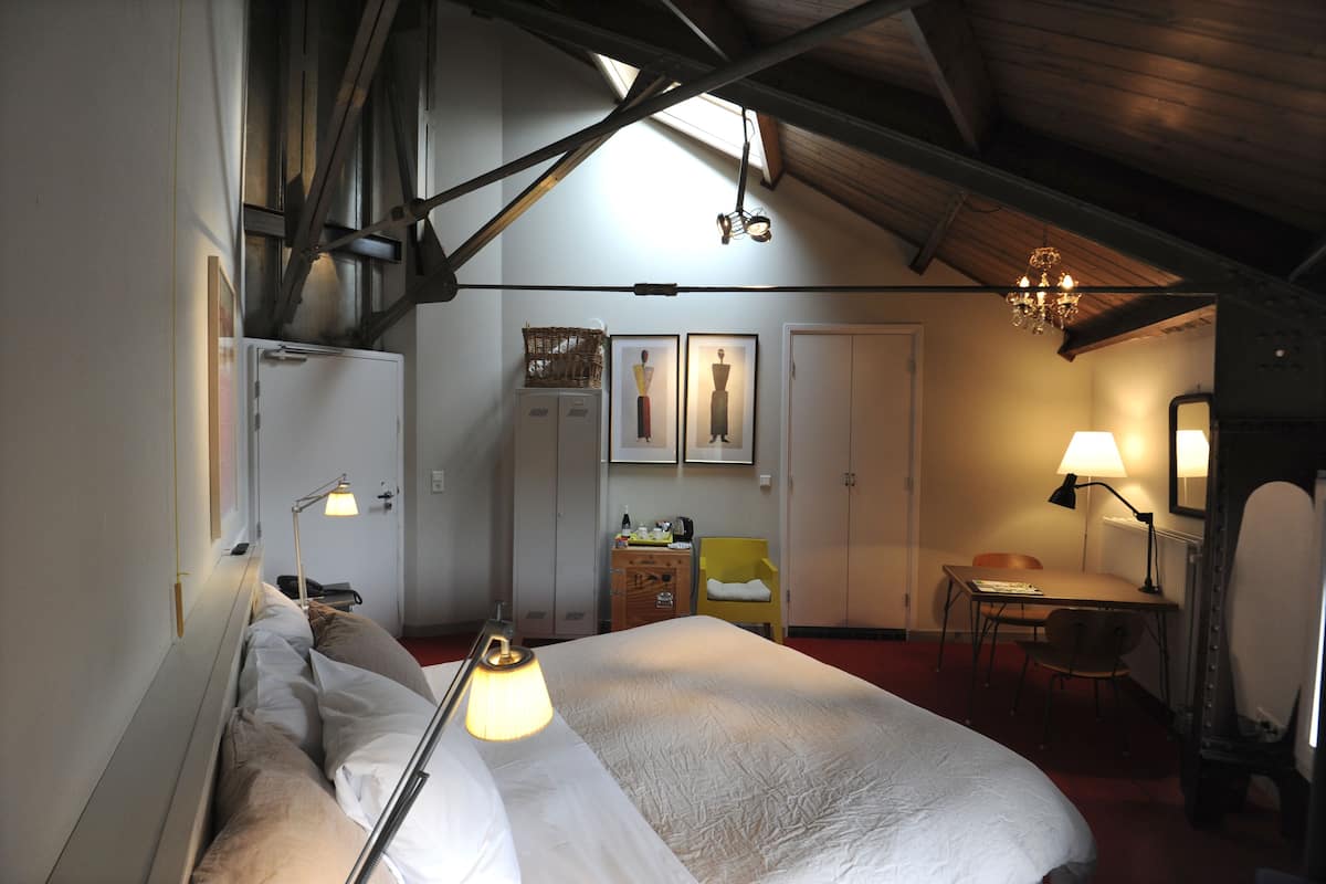 Habitación de hotel con cama, mesita de noche, lámpara, silla, escritorio y una pequeña mesa con lámpara. Se ve una ventana con vistas al exterior, y un candelabro colgando del techo.