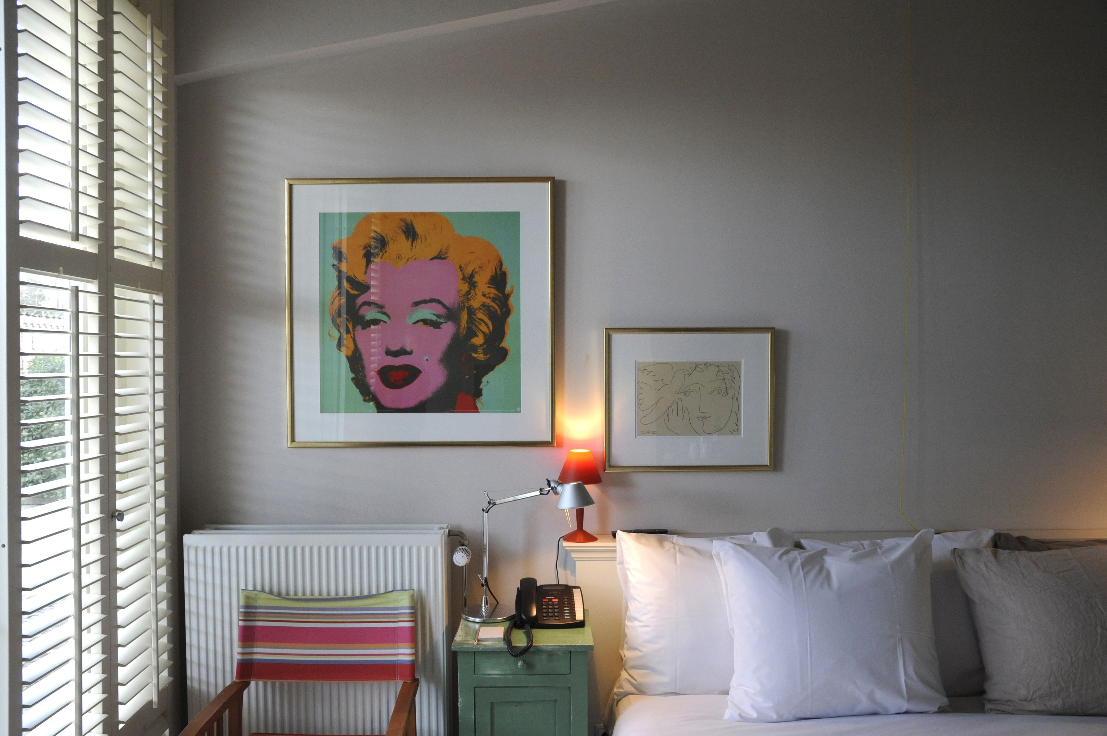 Un dormitorio con una cama, una mesita de noche con lámpara, un radiador y dos cuadros en la pared.