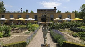 Un edificio con una estatua en primer plano, mesas al aire libre con sombrillas y un jardín con diversas plantas.