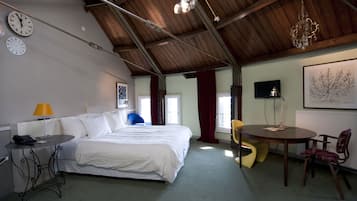 A spacious room with a large bed, a dining table, and a chandelier. There are clocks on the wall and a television on the wall.