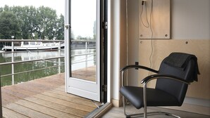 Una habitación moderna con vista a un río, una terraza de madera, una silla negra y una lámpara fijada a la pared.