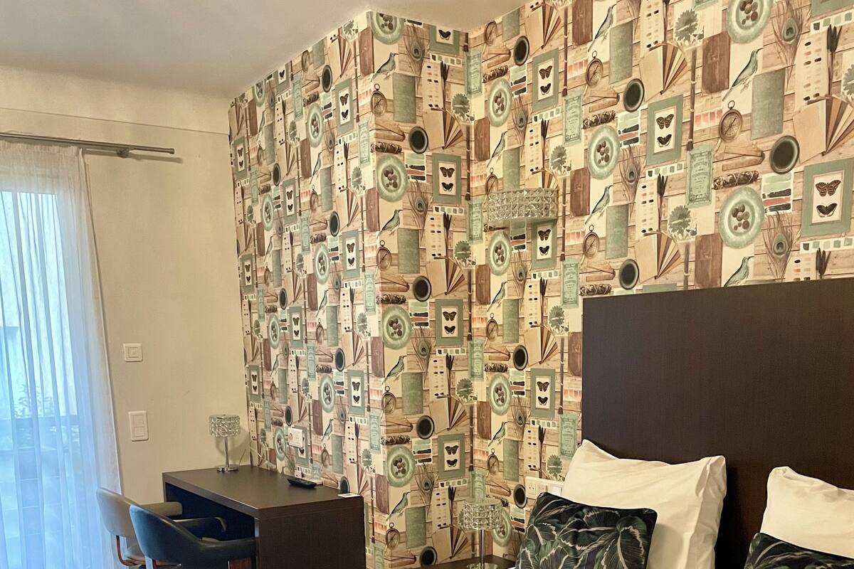 A hotel room with a patterned wallpaper, a bed with floral bedding, a desk, and a chair.