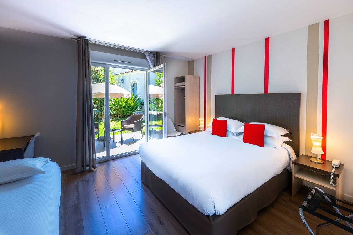 A modern hotel room with a large bed, a desk, and a chair. There is a sliding glass door leading to an outdoor area with a chair and plants.