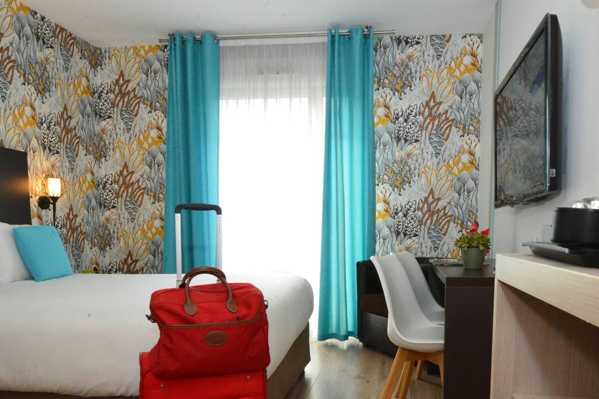 A hotel room with a bed, a desk, a chair, a red suitcase, and a wall-mounted television.