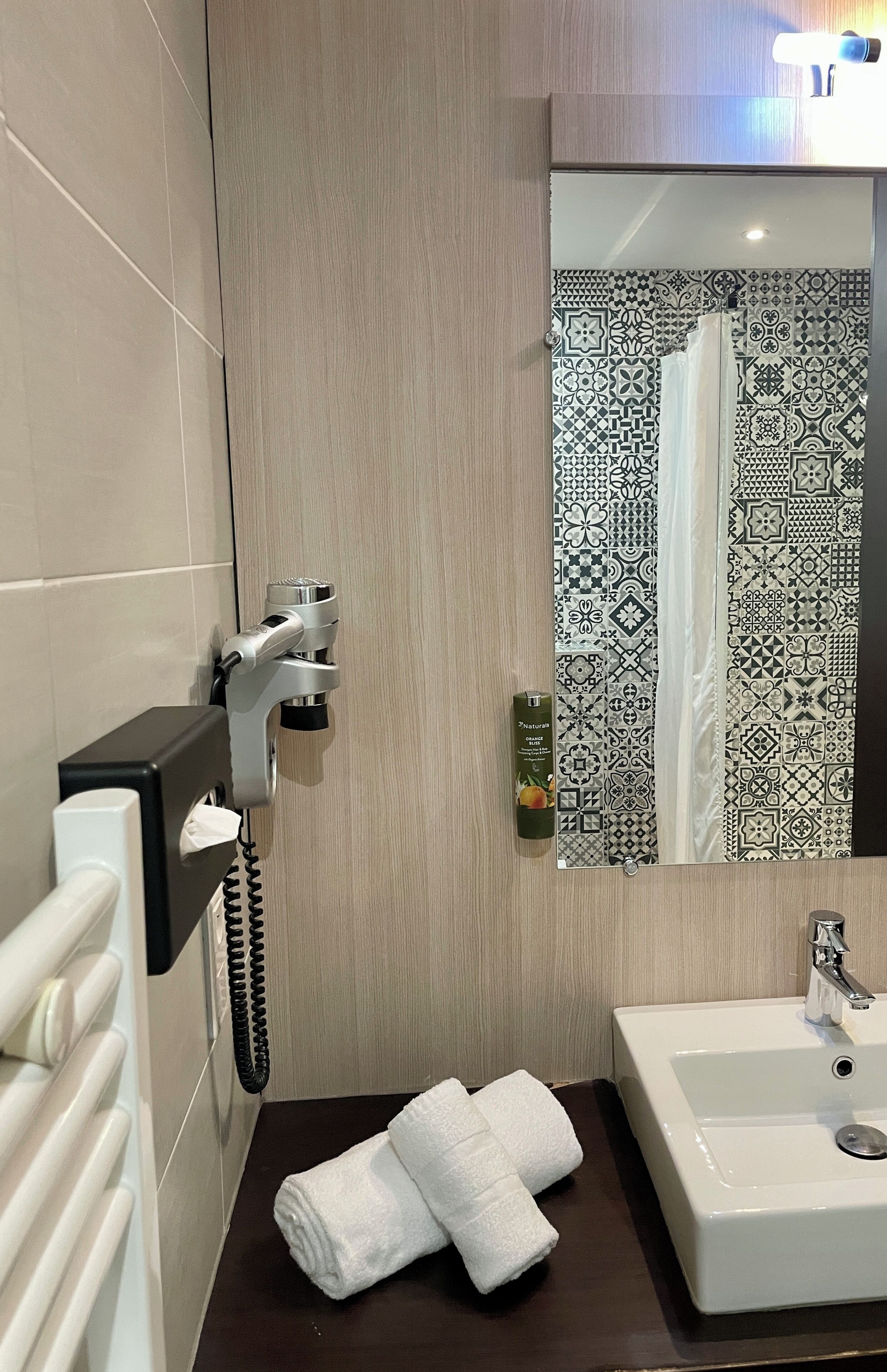 A bathroom with a hand dryer, a towel dispenser, a sink, and a mirror reflecting a shower curtain.