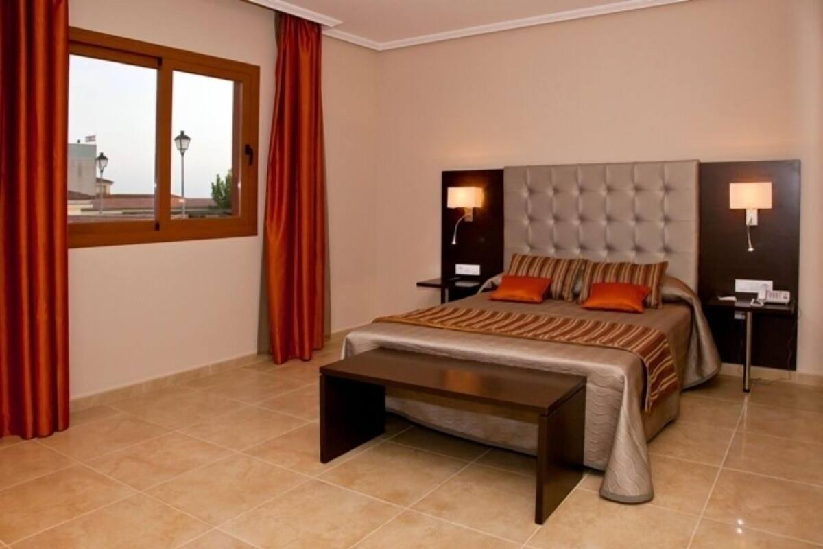 A modern hotel room with a large bed, two bedside lamps, a nightstand, and a window with curtains.
