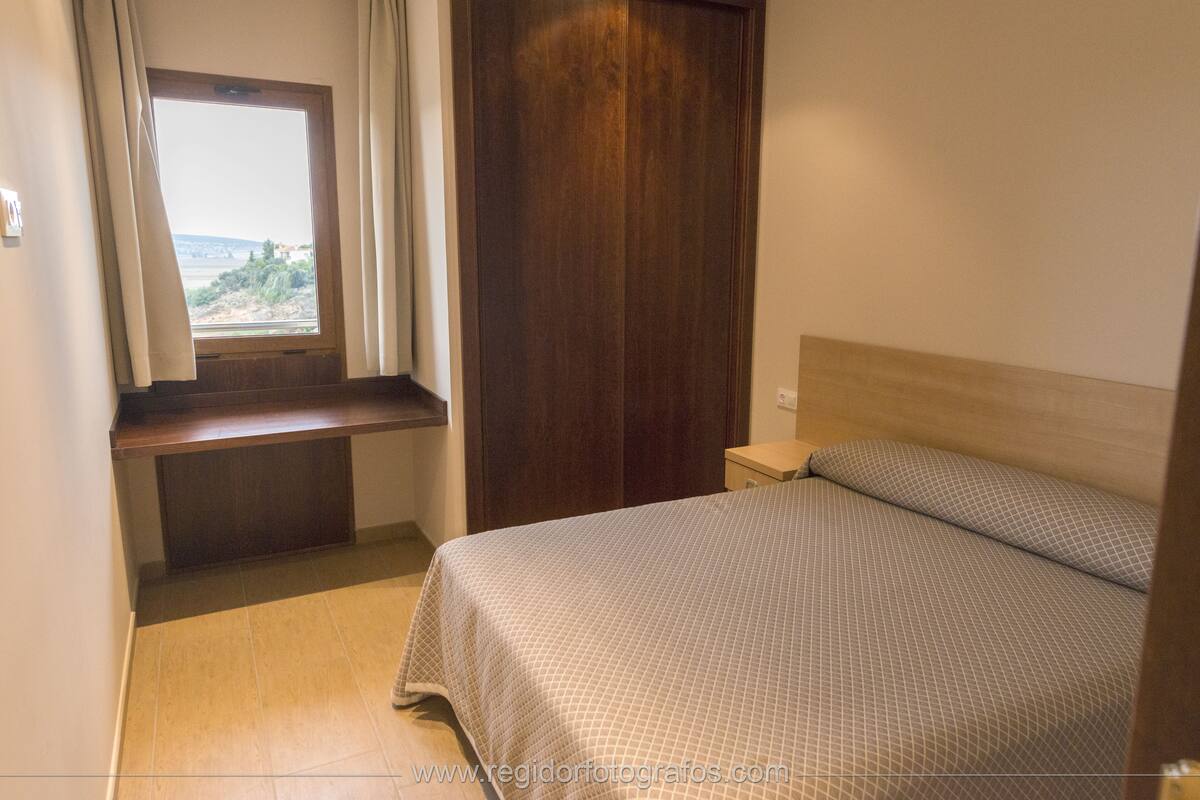 A bedroom with a bed, a window with curtains, a wooden wardrobe, and a bedside table.