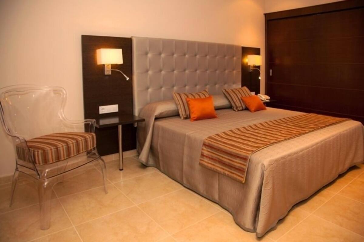 A bedroom with a large bed, a nightstand, a chair, and a wall-mounted lamp.