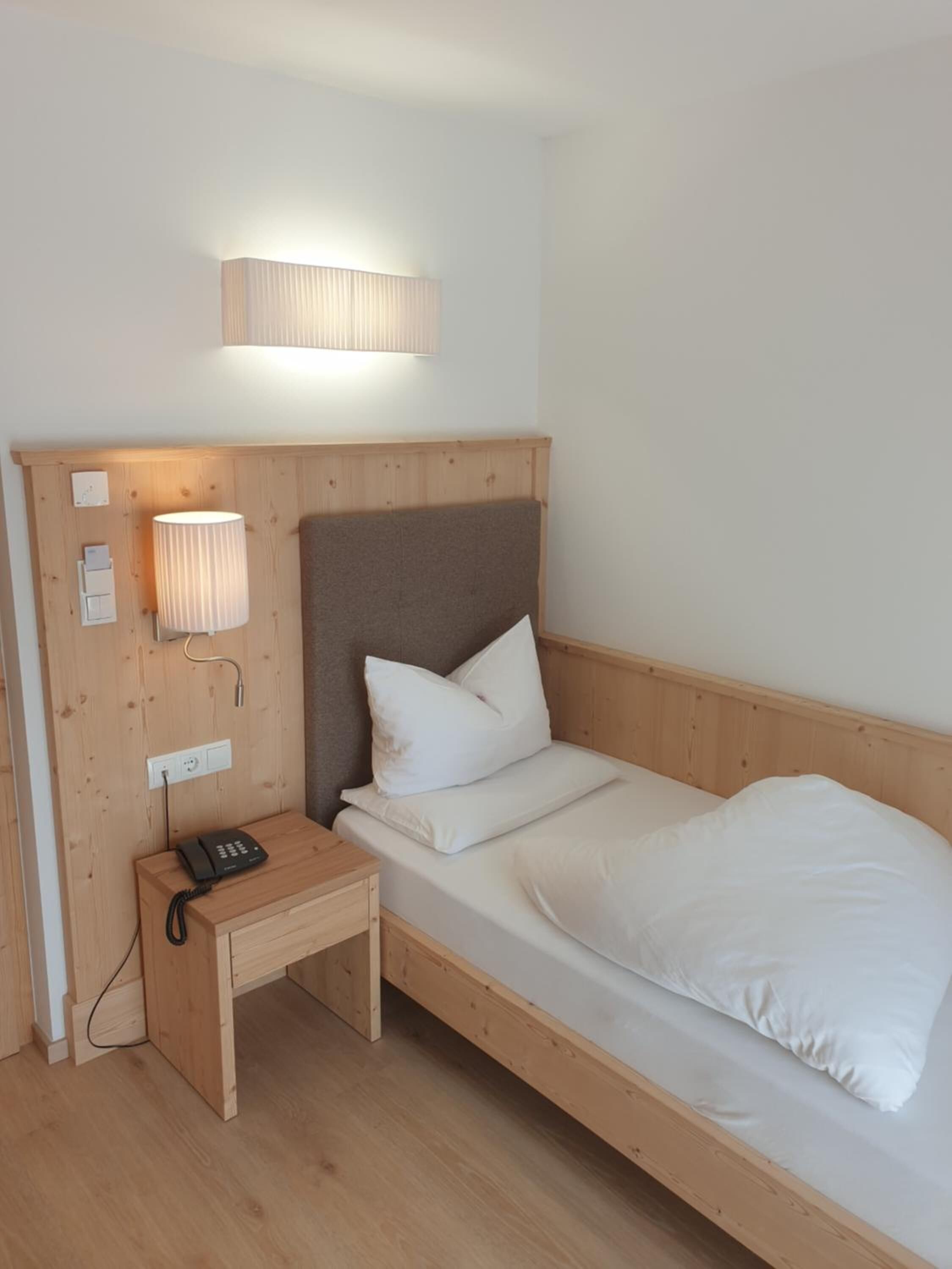 Une chambre avec un lit simple, une tête de lit en bois, une table de chevet avec une lampe et un téléphone.