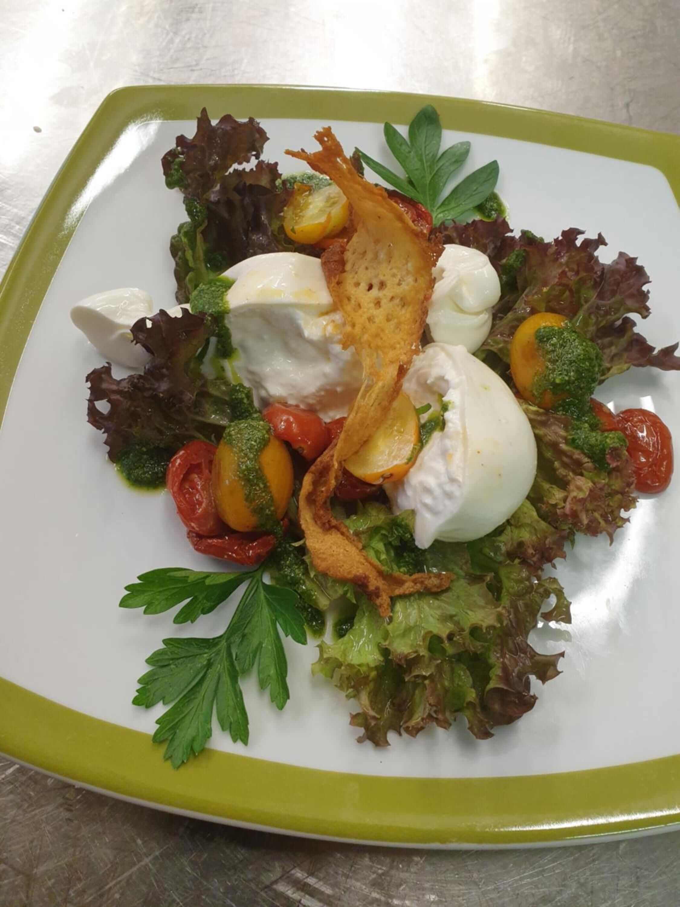 A square plate with a green rim containing a salad with lettuce, tomatoes, a poached egg, and a drizzle of green sauce.