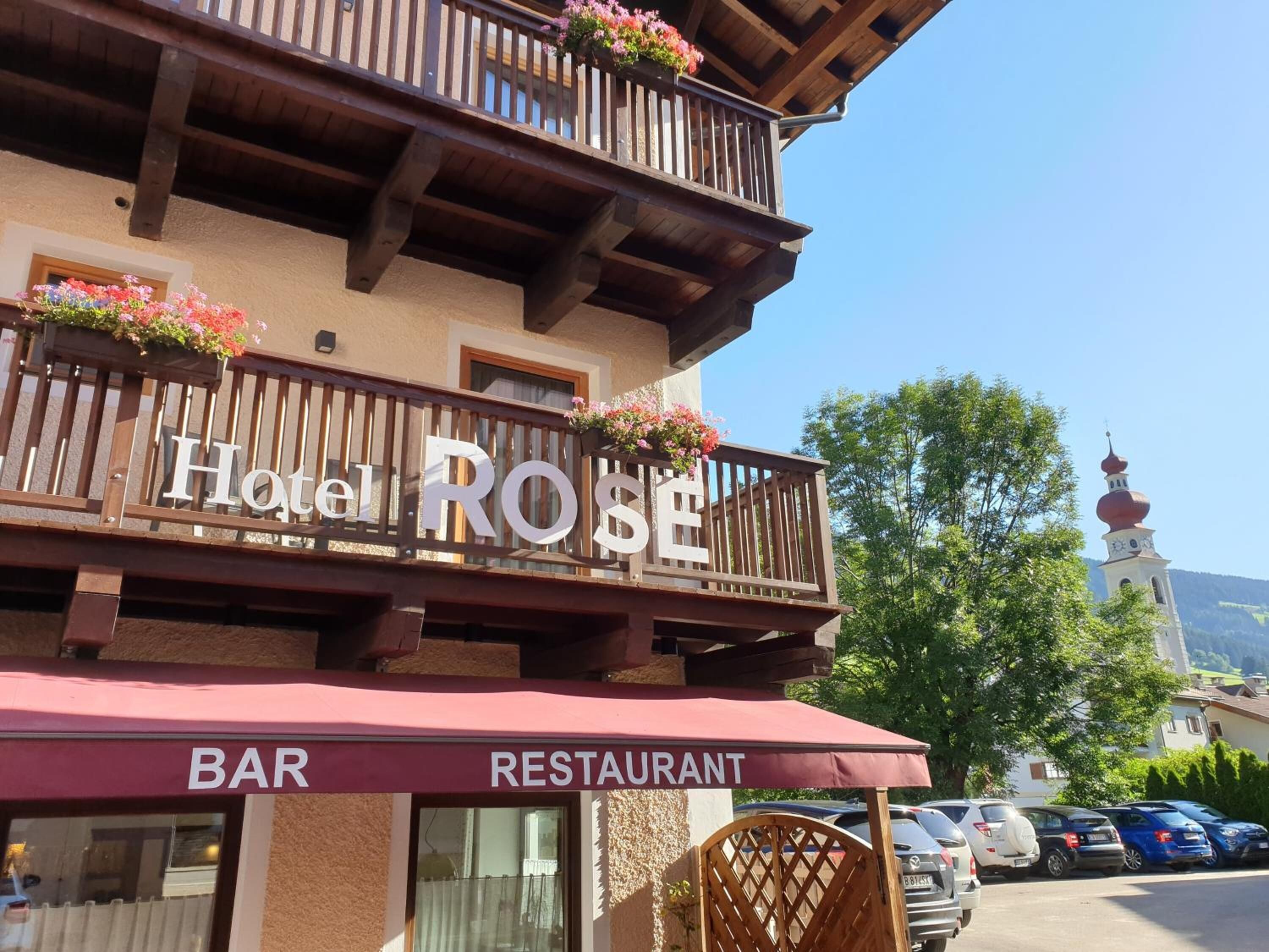 Hotel Rose con letrero de bar y restaurante, balcones de madera con jardineras y vista a una iglesia con cúpula roja.