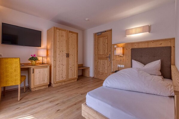 A hotel room with a bed, wooden wardrobe, desk, chair, and a TV mounted on the wall. - Hotel Rose (Villabassa)