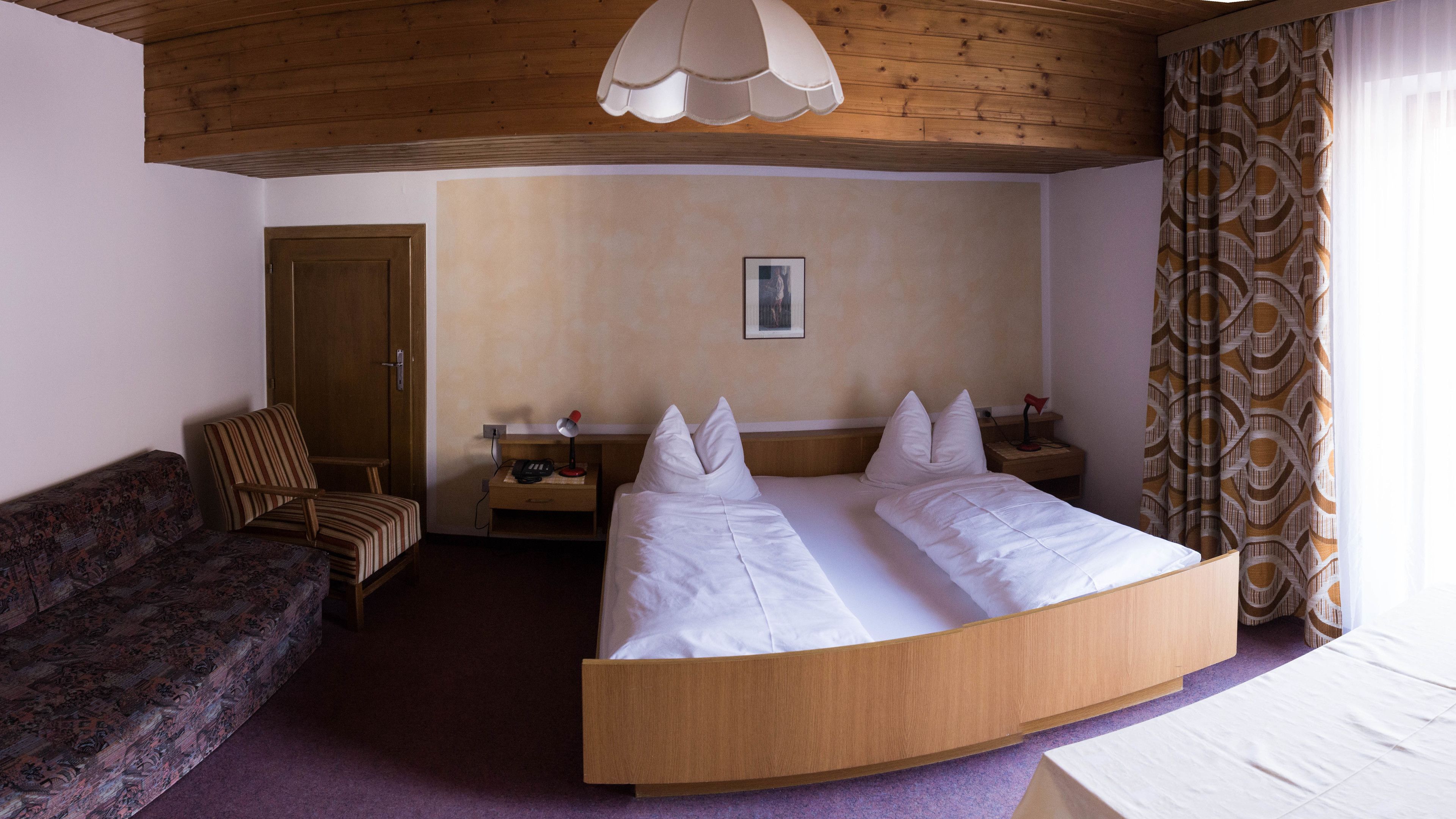 Una habitación con una cama de madera curvada, techo de madera, cortina estampada, mesita de noche de madera, un escritorio con lámpara, un sofá con diseño y una puerta.