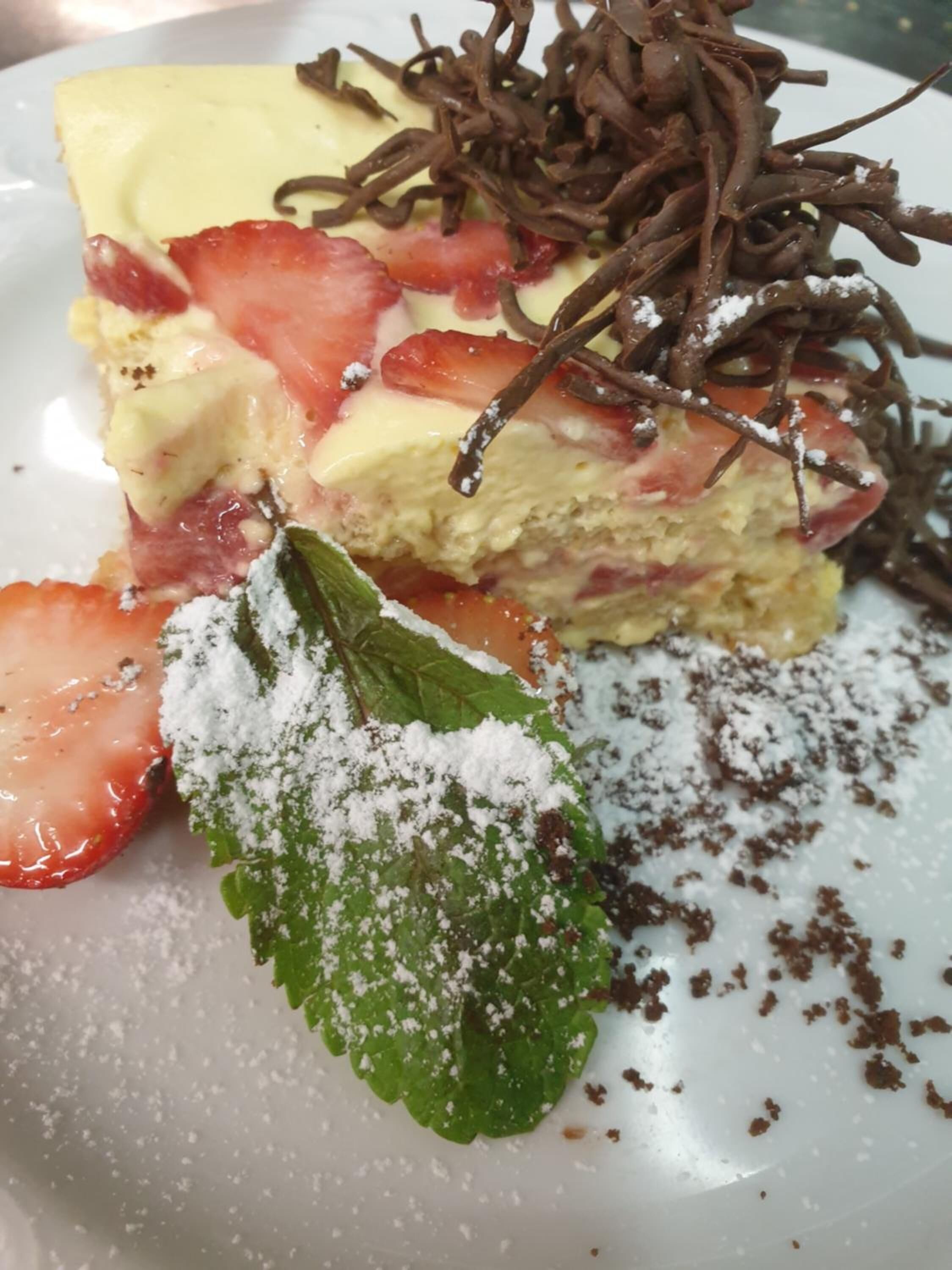 Un plat de postre amb una porció de pastís coronada amb maduixes i escates de xocolata, acompanyat d'una fulla de menta i sucre glas.