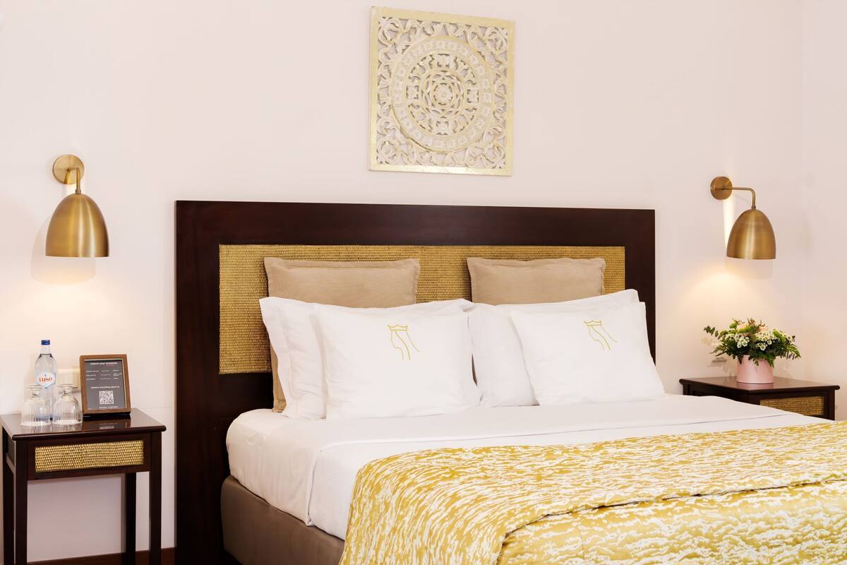 Un lit bien fait, avec du linge de lit blanc et une couvre-lit jaune à motifs, flanqué de deux tables de chevet avec des lampes, d’une œuvre d’art accrochée au mur et d’une petite plante en pot.