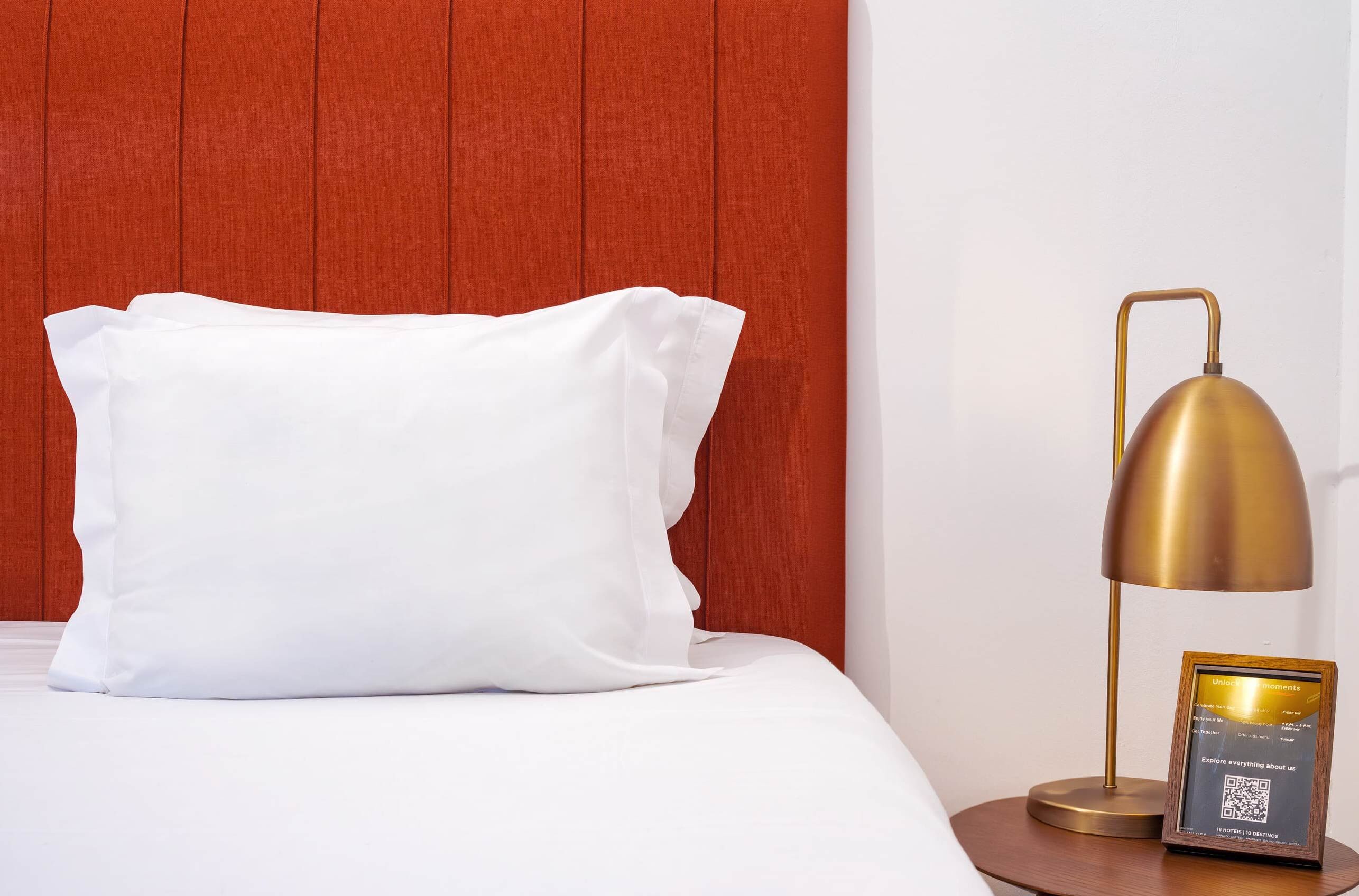 A neatly made bed with a white pillow and a gold lamp beside it.