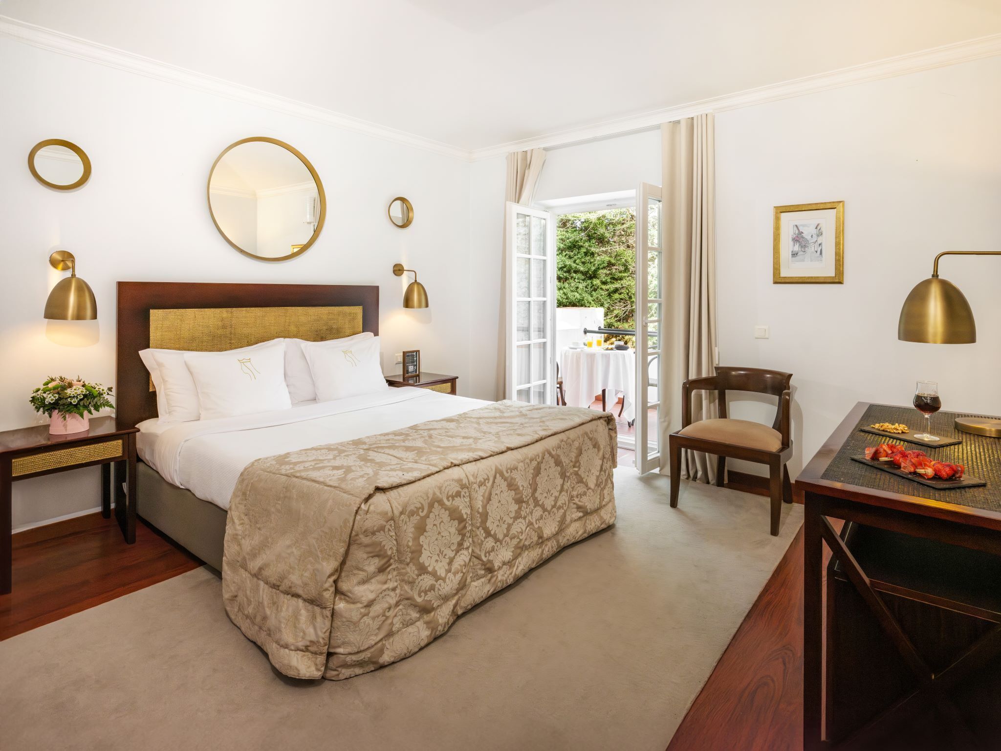 A hotel room with a large bed, two bedside tables with lamps, a desk with a chair, and a balcony with a table and chairs.