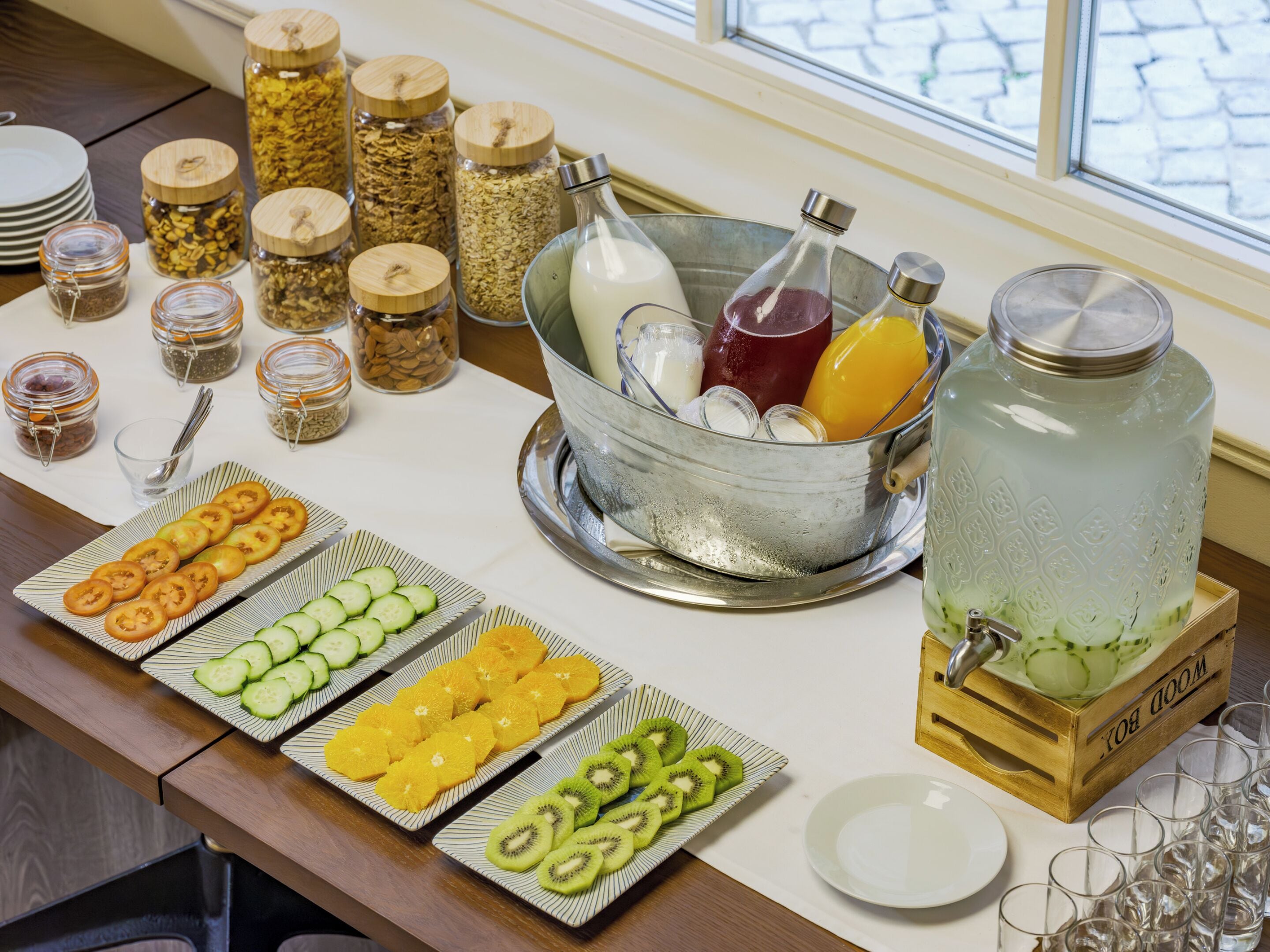 Una mesa de buffet con diversos alimentos y bebidas, como frutas cortadas, cereales y un gran jarro de agua.