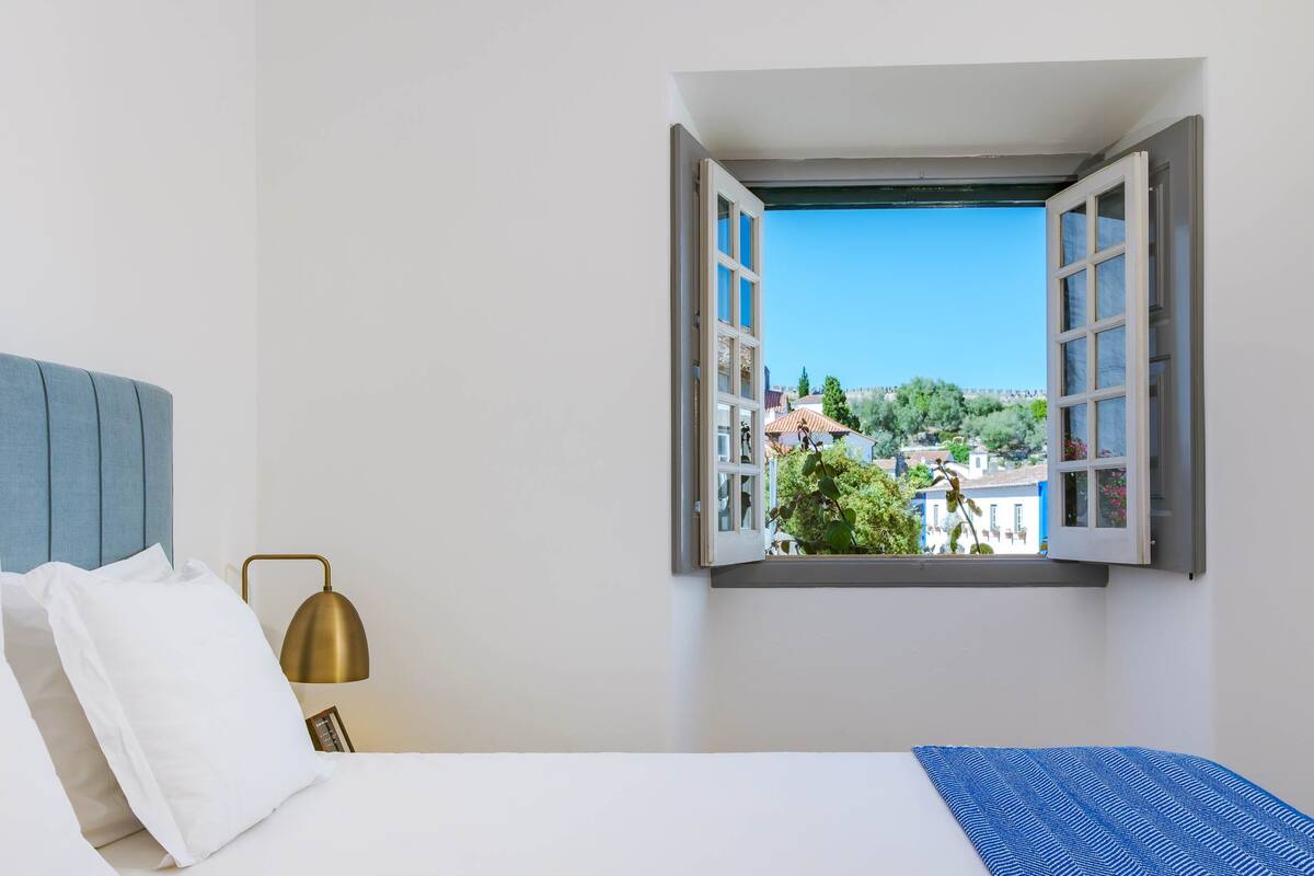 Une chambre à coucher avec un lit, une table de chevet, une lampe et une fenêtre dont le volet est ouvert.