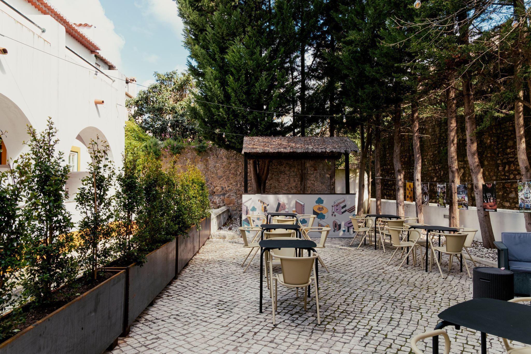 Un espace café en plein air, avec des tables et des chaises, entouré d’arbres et d’un mur de pierre.