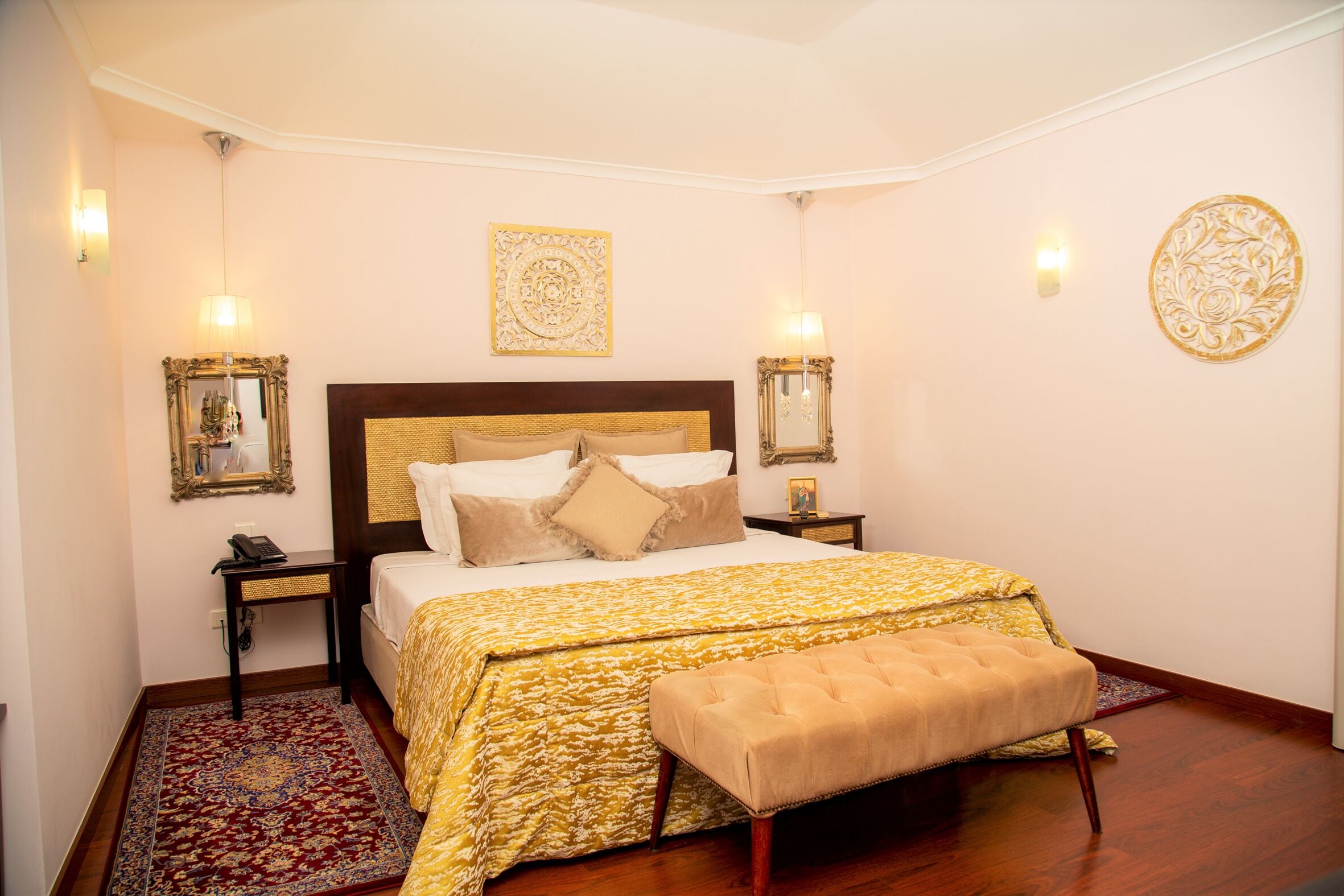 A bedroom with a large bed, a bench, a patterned rug, and decorative wall sconces.