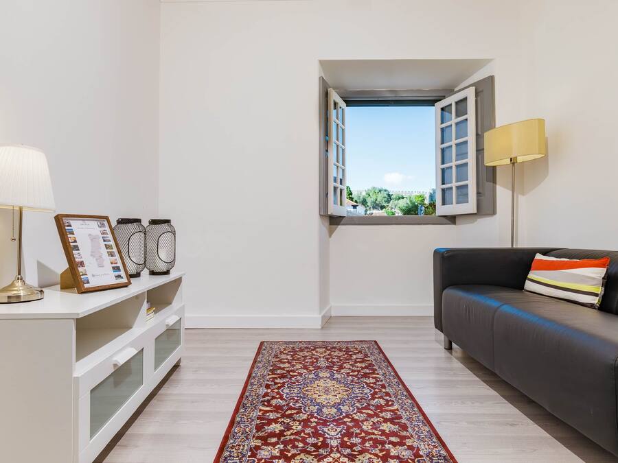 A modern living room with a sofa, a lamp, a small table with a picture frame, a patterned rug, and a window with an open shutter.