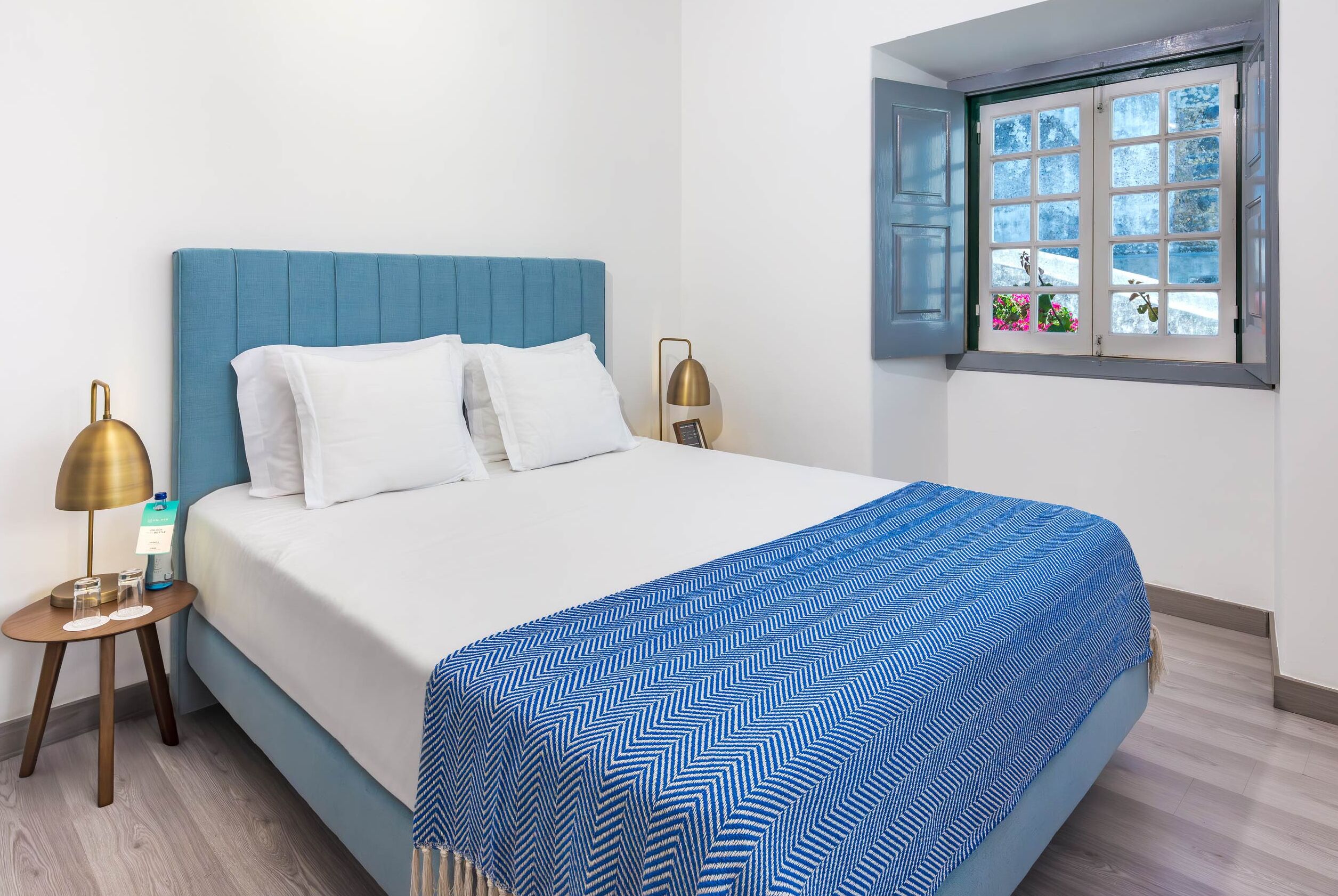 A neatly made bed with a blue headboard, white pillows, and a blue and white patterned bedspread. There are two bedside tables with lamps and a small picture frame on one. A window with a view of snow and flowers is visible.