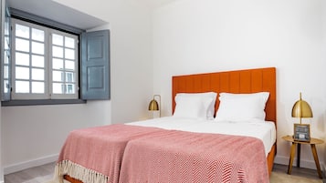 A bedroom with a bed, a window with blue shutters, and a bedside table with a lamp.