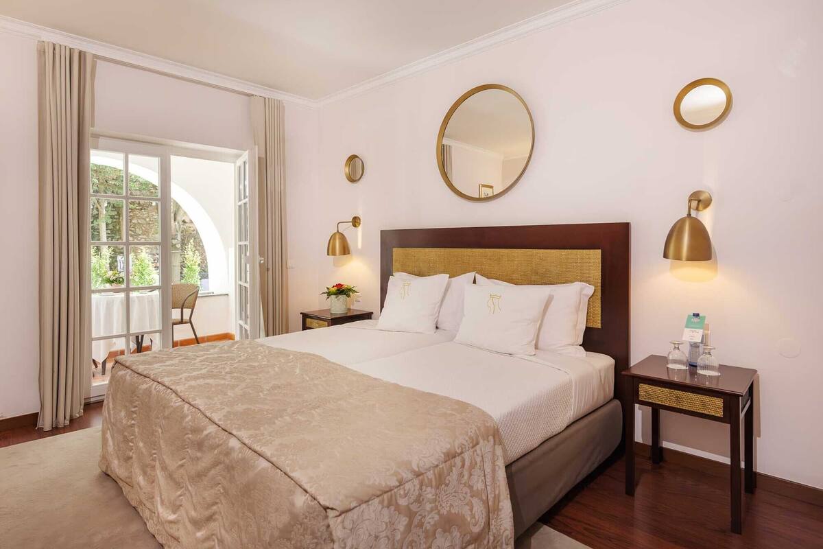Une chambre d’hôtel avec un grand lit, une petite table avec une lampe, un miroir au mur et une vue sur l’extérieur par une fenêtre.