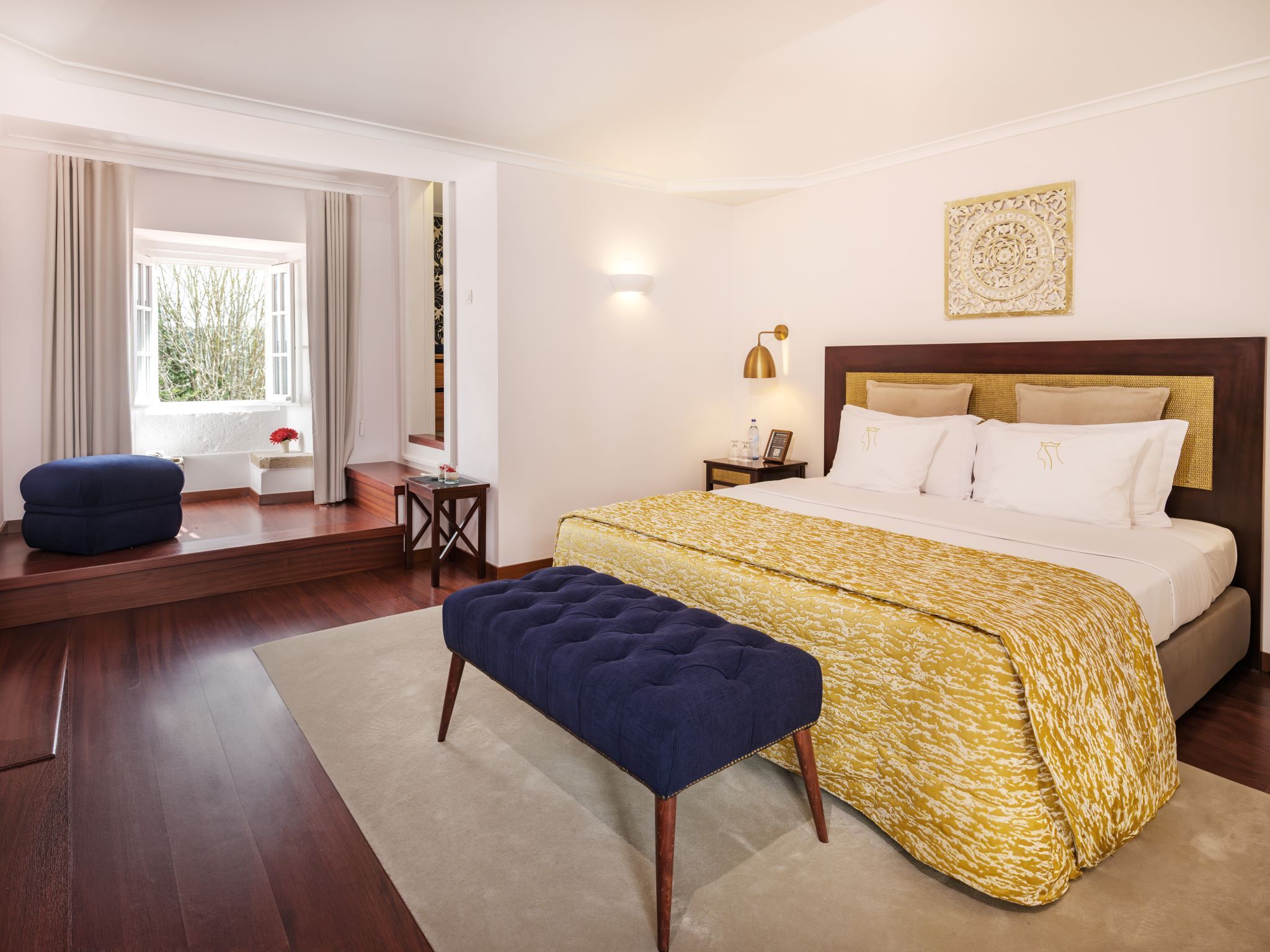 A bedroom with a large bed, a bench, a desk, and a window with curtains.