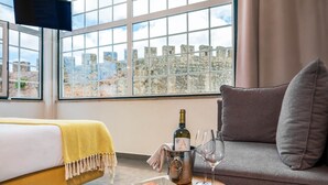 Hotelkamer met een bed, een grijze bank, een raam met uitzicht op stenen muren, een fles wijn en een fruitschaal.