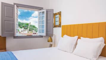 Habitación de hotel con cama, ventana con persianas abiertas, lámpara de noche y un cuadro en la pared.