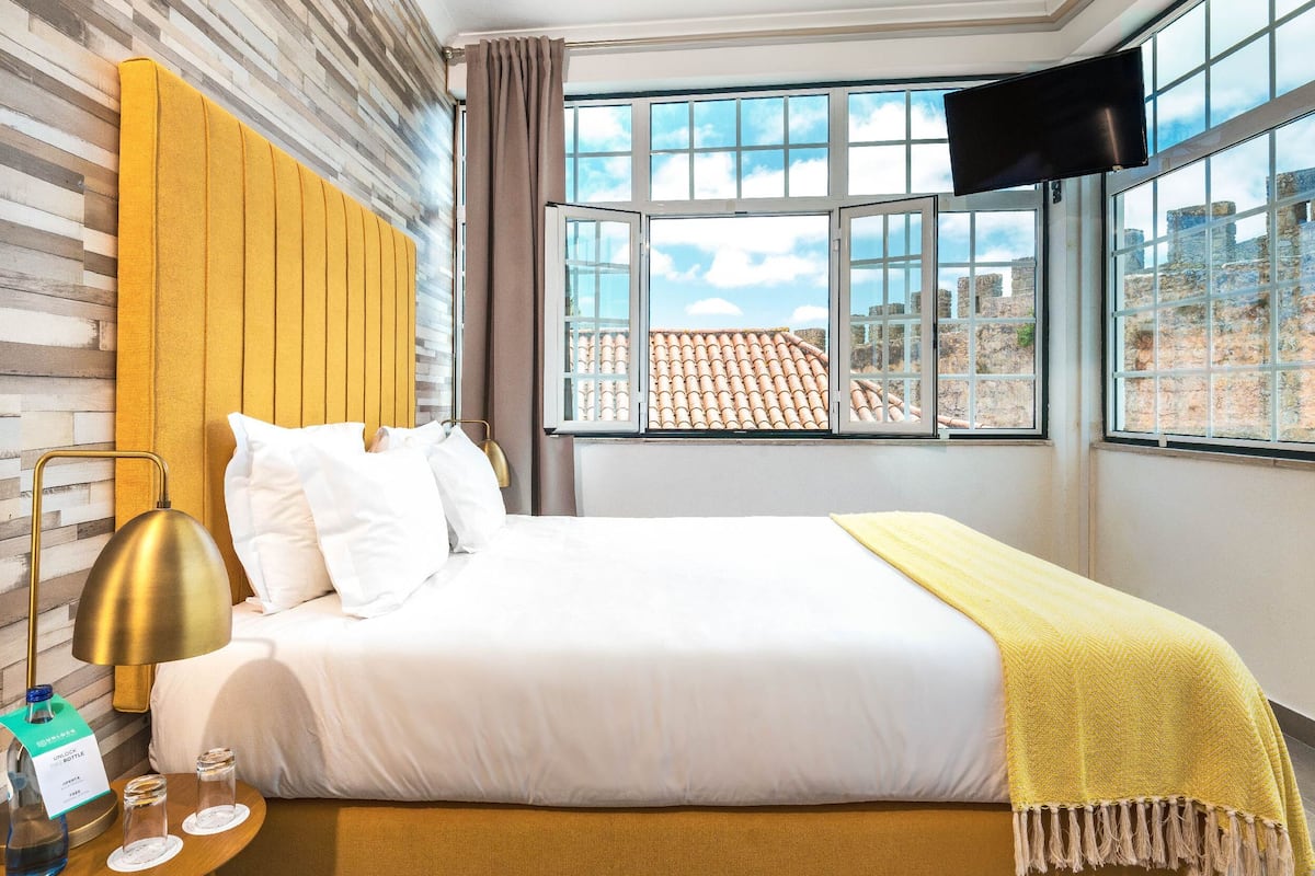 Une chambre d’hôtel avec un lit, un téléviseur fixé au mur, une fenêtre donnant sur des immeubles et une table de chevet avec une lampe et des lunettes.